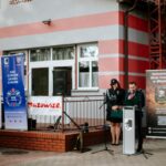 Grupa ludzi w mundurach strażackich stoi przed budynkiem z czerwono-szarymi pasami. Jedna osoba przemawia do mikrofonu. Obok znajdują się banery z napisem Mazowsze.