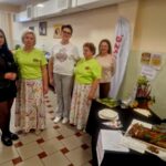 Na zdjęciu znajduje się pięć osób w jasnym pomieszczeniu. Kobiety mają na sobie kolorowe ubrania i stoją obok stołu. Na stole leży jedzenie i dekoracje, w tle widać plakaty i flagę.