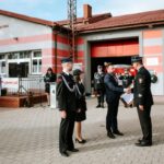 Grupa strażaków stoi przed remizą, odbierając nagrodę. Budynek jest szaro-czerwony, z otwartymi drzwiami garażu. W tle widoczne są pojazdy strażackie.