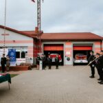 Na zdjęciu remiza strażacka z czerwonymi drzwiami i flagami. Strażacy w mundurach stoją przodem na placu. Tło z chmurami.