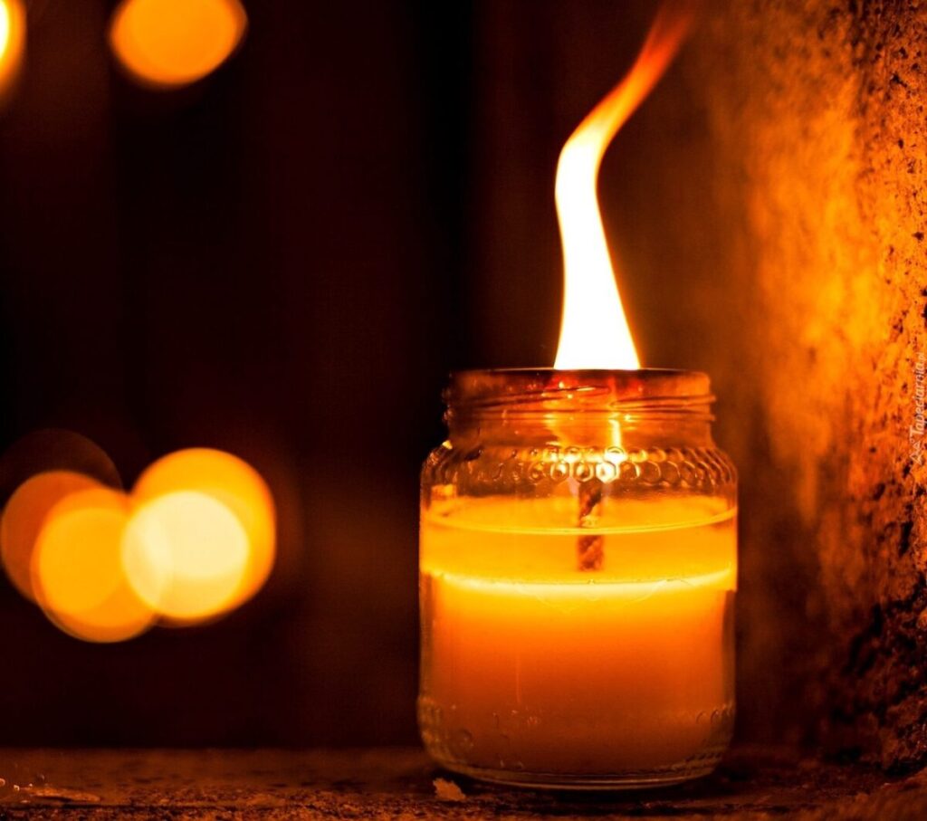 Świeca w szklanym słoiku płonie jasnym płomieniem. Dominują ciepłe pomarańczowe barwy. Tło jest ciemne z miękkimi konturami.