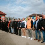 Grupa osób w strojach galowych i kurtkach stoi na placu brukowanym. Tło stanowi budynek z czerwonymi drzwiami i flagi. Niebo jest pochmurne.