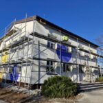 Budynek z rusztowaniami, biały z niebieskimi i żółtymi elementami. Na rusztowaniu pracownik w zielonej kurtce. Tło to jasne, niebieskie niebo.