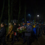 DSC_5469 Grupa ludzi maszeruje nocą w parku, niosąc wieńce. Ubrani w ciemne płaszcze i mundury, w tle drzewa i latarnie. Widoczne polskie flagi i symbolika patriotyczna.