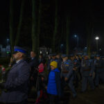 DSC_5473 Ludzie w mundurach i cywile uczestniczą w nocnej paradzie. Tło to oświetlona ulica z drzewami i budynkami. Dzieci w kolorowych ubraniach idą z dorosłymi.