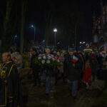 DSC_5479 Grupa ludzi maszeruje nocą, trzymając kwiaty i flagi. W tle oświetlony, ceglasty kościół. Otoczenie parku, ciemne kolory.