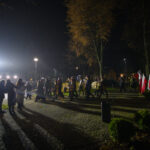 DSC_5484 Na zdjęciu nocą marsz ulicą, w tle polskie flagi. Tłum ludzi idzie oświetlony latarniami. Otoczenie parku, drzewa jesienią.