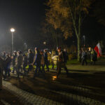 DSC_5489 Nocny marsz z flagą Polski i orkiestrą dętą idącą ulicą. W tle drzewa i latarnia. Uczestnicy w ciemnych ubraniach.