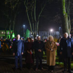 DSC_5495 Grupa ludzi stoi na placu nocą. W tle widoczne drzewa i budynki, oświetlone reflektorami. Uczestnicy trzymają flagi Polski.