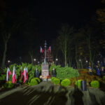 DSC_5502 Nocna uroczystość przy pomniku, otoczonym zielenią i flagami Polski. W tle park z drzewami i spacerującymi ludźmi.