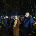 DSC_5504 Grupa ludzi stoi na zewnątrz nocą. Mają na sobie płaszcze, w tle drzewa i światła uliczne. Uczestniczą w wydarzeniu.