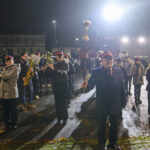 DSC_5506 Orkiestra marszowa w mundurach gra instrumenty na oświetlonym placu. Nocne tło z budynkami i samochodami. Dominują ciemne kolory.