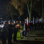 DSC_5517 Grupa umundurowanych ludzi stoi w nocnym parku, trzymając flagi i wieńce. Dominuje ciemna kolorystyka z oświetleniem lamp ulicznych. Tło stanowią drzewa i budynki.