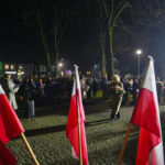 DSC_5520 Grupa ludzi na wieczorze, w tle ciemne niebo i drzewa. Flagi polskie dominują na pierwszym planie. Postać z kwiatami idzie przed tłum.