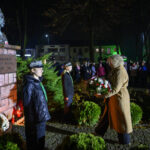 DSC_5523 Pomnik Józefa Piłsudskiego nocą, otoczony zielenią. Kobieta składa wieniec, przygląda się jej tłum w tle. Jasne oświetlenie.
