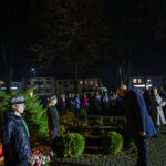 DSC_5534 Pomnik marszałka Piłsudskiego na tle parku nocą. Ludzie zgromadzeni w ciemnych ubraniach, wokół kwiaty i znicze. Tło ciemne z ciepłym oświetleniem.