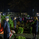 DSC_5537 Pomnik marszałka Piłsudskiego w parku nocą. Ludzie składają kwiaty, otoczenie oświetlone sztucznym światłem. Tło to ciemne drzewa i budynki.