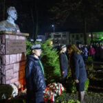 DSC_5540 Pomnik Piłsudskiego nocą, otoczony zielenią. Grupa ludzi w tle, światła i wieńce. Uroczystość upamiętniająca.