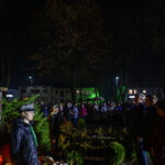 DSC_5549 Pomnik na tle tłumu w parku nocą. Osoby w mundurach stoją po lewej. Otoczenie oświetlone jest zielonym i niebieskim światłem.