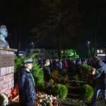 DSC_5556 Popiersie marszałka na postumencie z cegły otoczonego wieńcami. Nocna sceneria z ludźmi i zielonymi krzewami w tle. Oświetlone budynki i drzewa.