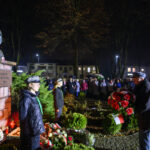 DSC_5565 Pomnik Piłsudskiego oświetlony nocą, otoczony przez ludzi z biało-czerwonymi kwiatami. Tło to ciemne niebo i drzewa. Wydarzenie upamiętnia ważną osobę.