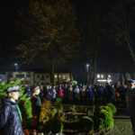 DSC_5572 Pomnik Marszałka Piłsudskiego nocą otoczony przez ludzi. Tło z drzewami, w oddali widoczne budynki. Oświetlenie białe i żółte.