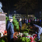 DSC_5578 Pomnik z popiersiem mężczyzny stoi w parku nocą. Wokół są ludzie, żołnierze i kwiaty w biało-czerwonych barwach. Za tłem widać budynki.