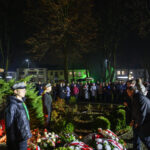 DSC_5587 Pomnik w parku podczas nocnej uroczystości. Ludzie zgromadzeni wokół, z kwiatami i flagami. Oświetlenie w odcieniach zieleni.