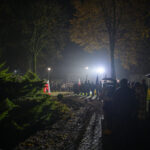 DSC_5594 Nocna scena z tłumem ludzi stojących wzdłuż oświetlonej ścieżki. W tle drzewa, światła, jesienne liście. Flaga Polski widoczna po lewej.