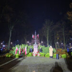 DSC_5596 Pomnik otoczony zielenią z polskimi flagami na tle nocnego parku. Ludzie stoją wokół, słuchając przemówienia. Ciepłe światła oświetlają scenę.