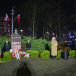 DSC_5599 Na nocnym zdjęciu widoczny jest pomnik z popiersiem na tle drzew, flagi Polski i grupa ludzi. Obok stoi osoba z mikrofonem, a w tle znajdują się oświetlone budynki. Dominuje ciemne, zielono-brązowe tło.