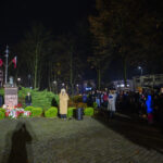 DSC_5600 Nocna uroczystość przy pomniku z flagami i kwiatami. Kobieta przemawia, ludzie zgromadzeni obok. Drzewa w tle i ceglana droga.