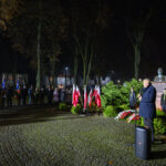 DSC_5601 Nocne zgromadzenie na placu, z mężczyzną przemawiającym przy mikrofonie. Liczne polskie flagi na tle zielonych krzewów. Pomnik i tłum ludzi w tle.