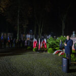 DSC_5602 Nocna uroczystość z uczestnikami, flagami polskimi i przemawiającym. Zielone krzewy z pomnikiem w tle. Oświetlenie jasne, ciepłe.