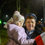 DSC_5611 Mężczyzna w mundurze trzyma dziecko w różowym ubraniu z czapką. Dziecko trzyma polską flagę. W tle nocne rozmyte światła i drzewa.