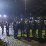 DSC_5612 Grupa ludzi w mundurach stoi w deszczu nocą. Po lewej muzycy z instrumentami, tło ciemne z drzewami i światłami. Mundury szaro-niebieskie.