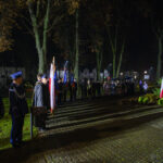 DSC_5613 Grupa ludzi w mundurach stoi w parku nocą. Widać biało-czerwone flagi Polski oraz drzewa i bloczy w tle. Teren jest oświetlony lampami ulicznymi.