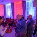 DSC_5622 Mężczyźni w mundurach z medalami, kobieta podaje wstążki z koszyka. Jasne zasłony w tle oświetlone na niebiesko i czerwono. Salę wypełnia grupa ludzi na czerwonych krzesłach.