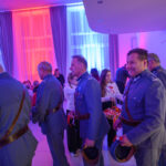 DSC_5624 Grupa ludzi w mundurach, trzymająca kapelusze. Tło oświetlone kolorowymi światłami: niebieskim i czerwonym. Na stole po lewej widoczne dekoracje.