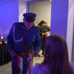 DSC_5626 Na obrazie starszy mężczyzna w mundurze wręcza flagę dziewczynce. Po lewej stół z jedzeniem, tło fioletowe. W tle inni ludzie z flagami.