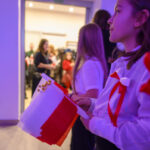 DSC_5629 Grupa osób zebrana w jasnym pomieszczeniu trzyma biało-czerwone flagi. Ubrani w białe koszule z czerwonymi akcentami. Światło tworzy purpurowy ton.