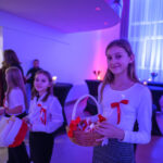 DSC_5630 Na zdjęciu widać trzy dziewczynki w białych bluzkach z czerwonymi wstążkami, trzymają koszyki. Tło jest jasno oświetlone na niebiesko i różowo. W tle widać ludzi oraz zasłony okienne.