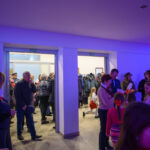 DSC_5636 Na zdjęciu widać grupę ludzi w jasnym pomieszczeniu. W tle płaszcze, a ściany są oświetlone na niebiesko i fioletowo. Niektórzy trzymają polskie flagi.