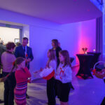 DSC_5637 Grupa ludzi stoi w jasnym pomieszczeniu. Młode dziewczyny trzymają flagi i koszyk. Tło jest niebieskie z różowym światłem.