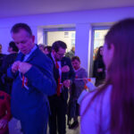 DSC_5642 Grupa ludzi w jasnym pomieszczeniu ozdabia siebie biało-czerwonymi wstążkami. Fioletowe oświetlenie dodaje nastroju. W tle inne osoby i stół z jedzeniem.