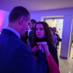 DSC_5653 Na zdjęciu widać mężczyznę przypinającego kotylion kobiecie. Dominują odcienie niebieskiego i fioletowego. Tło to eleganckie wnętrze z ludźmi.