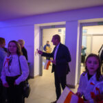 DSC_5655 Grupa osób w eleganckich strojach, trzyma biało-czerwone flagi. Tło oświetlone na fioletowo. W tle stół z przekąskami.