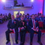 DSC_5673 Grupa ludzi w eleganckich strojach siedzi w sali oświetlonej na niebiesko i różowo. Trzymają flagi Polski. Tło to balkon z osobami obserwującymi.