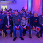 DSC_5678 Grupa ludzi siedzi w formalnej sali, ubrana elegancko z biało-czerwonymi kotylionami. Ściany oświetlone są na niebiesko i czerwono. Niektórzy trzymają polskie flagi.