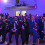 DSC_5685 Grupa ludzi siedzi w kilku rzędach na uroczystości. Mają biało-czerwone kokardy i flagi Polski. Tło oświetlone na niebiesko i różowo.